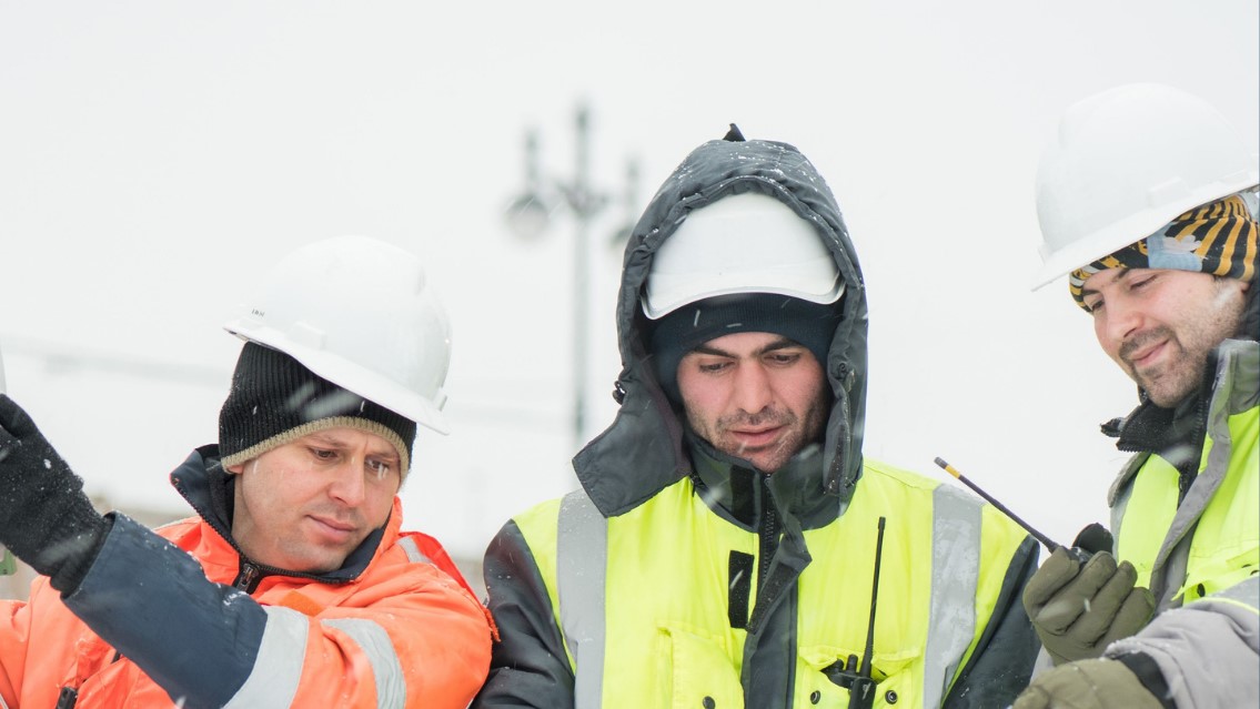 : Blog banner with image of three workers in hi-vis jackets, warm clothing, gloves, and helmets in the snow reading from a document. The text says “Cold Weather Safety Gear For Outdoor Workers”