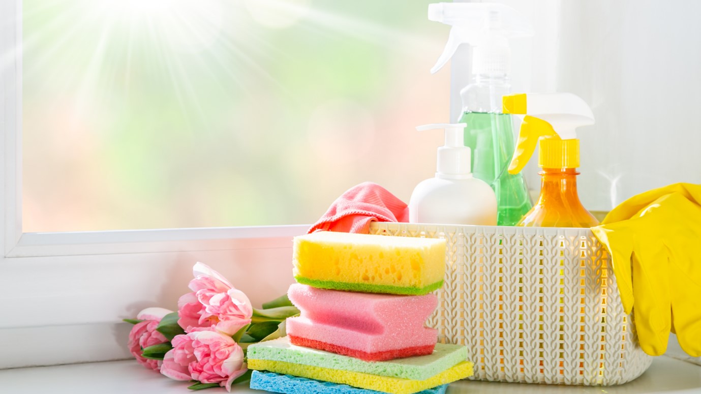 Image of cleaning products including sponges, cleaning spray and rubber gloves next to a sunny spring window.