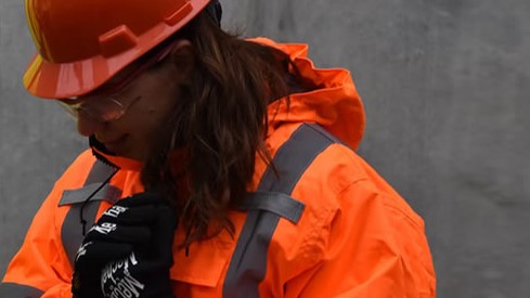 A woman wearing a helmet, PPE goggles, hi-vis jacket and protective gloves